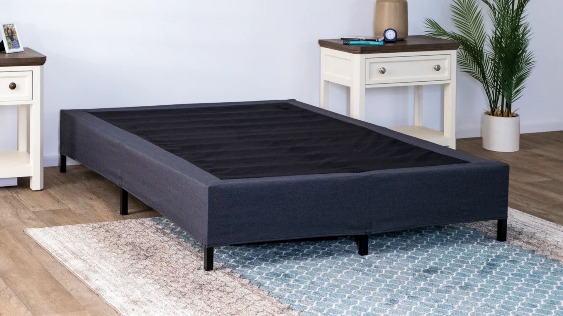 Modern bedroom with charcoal bed frame, white nightstand, geometric rug, and potted plant.